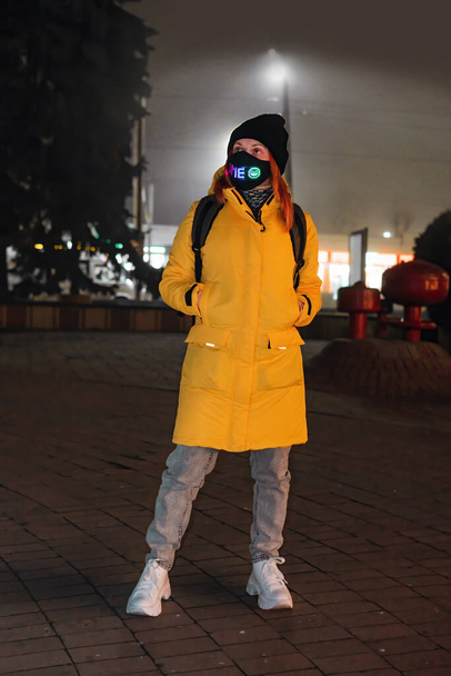 Uma menina vestida com um casaco de inverno amarelo fica em uma rua da cidade à noite. Há uma máscara no rosto. Quarentena e conceito de caminhada. Foto vertical - Foto, Imagem