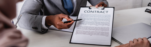 обрізаний вид афроамериканського бізнесмена, який вказує ручкою за контрактом біля ділових партнерів на розмитому фоні, банер
 - Фото, зображення
