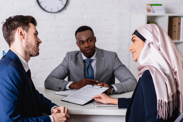 smiling arabian businesswoman pointing with hand at contract during discussion with multiethnic business partners, blurred background - Photo, Image