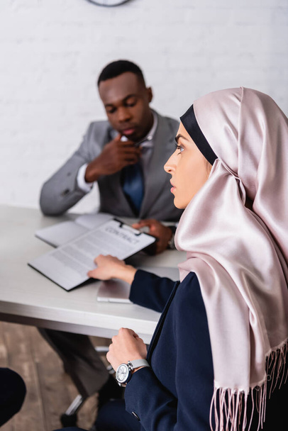арабская предпринимательница, указывающая рукой на контракт рядом с африканским американским бизнес-партнером на размытом фоне - Фото, изображение