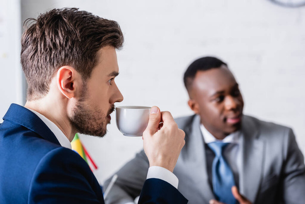 interpreter drinking coffee near african american business partner on blurred background - Photo, Image