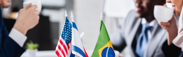 selektiver Fokus amerikanischer, brasilianischer und US-amerikanischer Flaggen in der Nähe multikultureller Geschäftsleute, die Kaffee auf verschwommenem Hintergrund trinken, Teilsicht, Banner - Foto, Bild