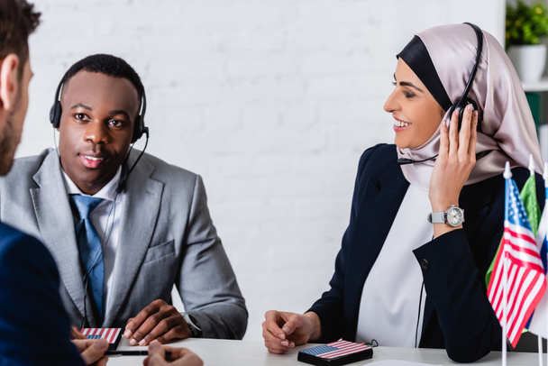 glimlachende Arabische en Afrikaanse Amerikaanse zakenpartners in headsets near interpreter op wazige voorgrond - Foto, afbeelding