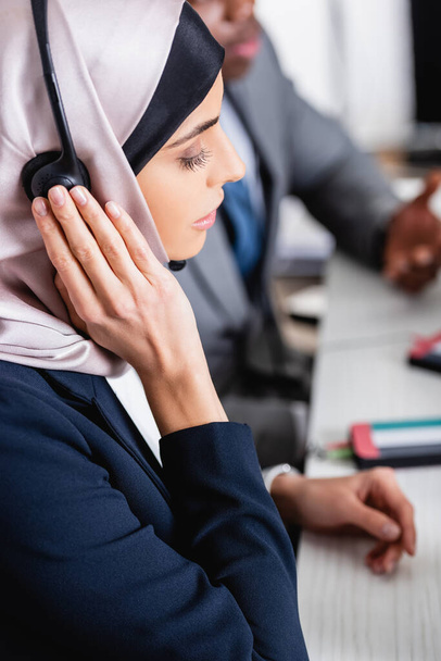 arabian businesswoman touching headset near african american business partner of blurred background - 写真・画像