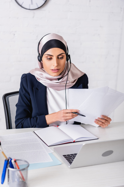 arabian interpreter in headset working with documents near blank notebook and laptop, blurred foreground - Φωτογραφία, εικόνα