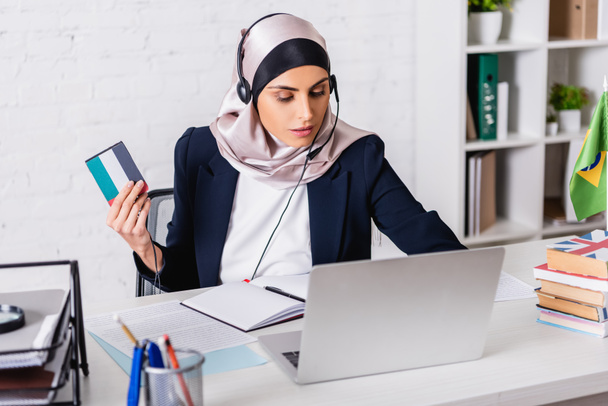 arabský tlumočník ve sluchátkách s digitálním překladačem při práci na notebooku v blízkosti slovníků, rozmazané popředí - Fotografie, Obrázek