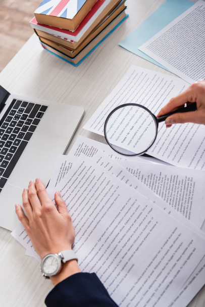partial view of translator holding magnifier above documents near laptop and dictionaries of foreign languages - Photo, Image