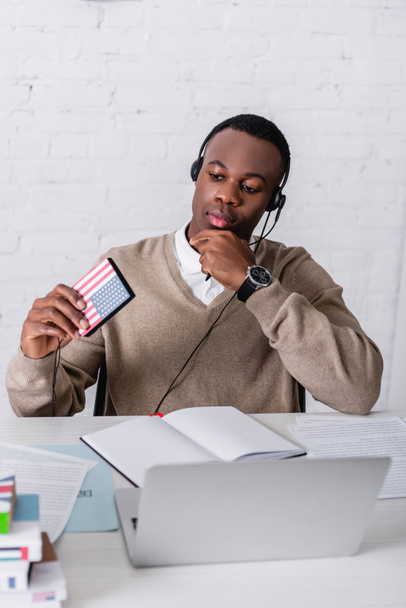 doordachte Afrikaans-Amerikaanse tolk in headset met digitale vertaler met Amerikaanse vlag embleem in de buurt van notebook en laptop op wazig voorgrond - Foto, afbeelding