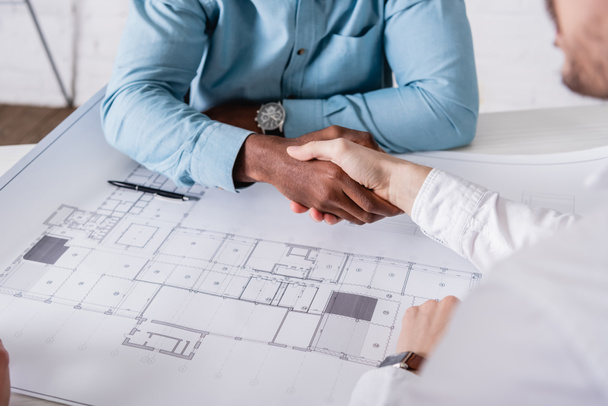 cropped view of african american businessman shaking hands with business partner near blueprint, blurred foreground - Photo, Image