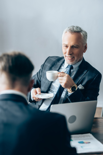 Inversor positivo sosteniendo taza de café cerca de portátil y hombre de negocios en primer plano borrosa  - Foto, Imagen