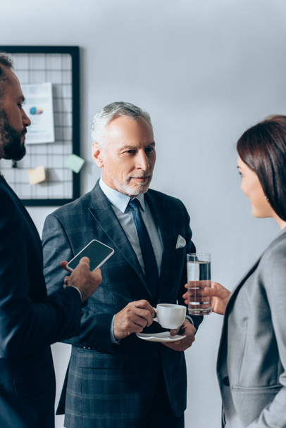 Businesswoman with glass of water standing near businessmen with coffee and smartphone in office  - Foto, Imagen