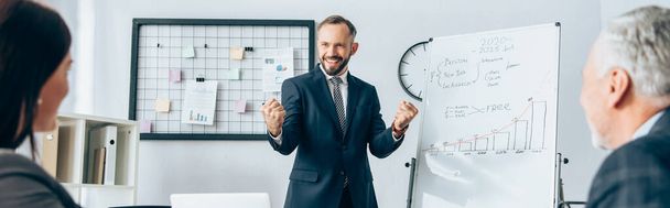 Cheerful businessman showing yeah gesture near flipchart and partners on blurred foreground in office, banner  - Foto, Bild