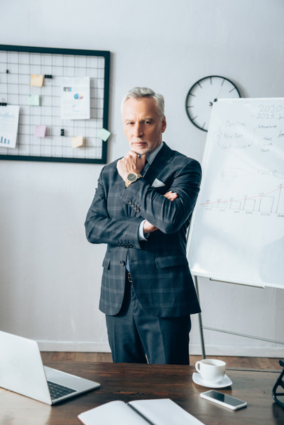 Confident businessman looking at camera near gadgets and notebook on blurred foreground  - Foto, Bild