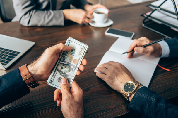 Ausgeschnittene Ansicht eines Geschäftsmannes mit Bargeld in der Nähe von Kollegen mit Notizbuch und Kaffee auf verschwommenem Hintergrund  - Foto, Bild