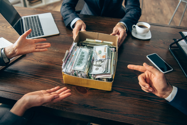 Cropped view of businesspeople pointing at box with money near colleague and devices on blurred foreground  - Photo, Image