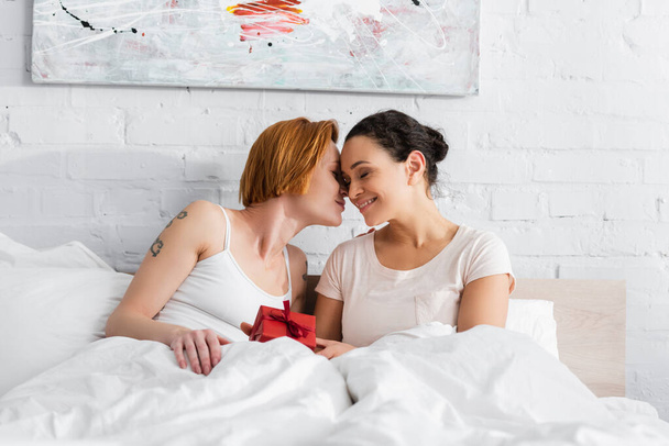 redhead lesbian woman kissing african american girlfriend holding valentines day gift in bed - Foto, imagen