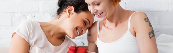 happy lesbian interracial girlfriends smiling near gift box on valentines day in bedroom, banner - Photo, Image