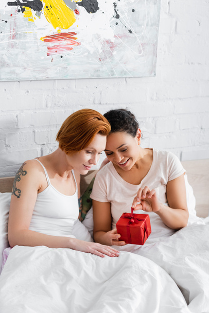 happy african american woman opening gift box near redhead lesbian girlfriend in bed - Fotografie, Obrázek