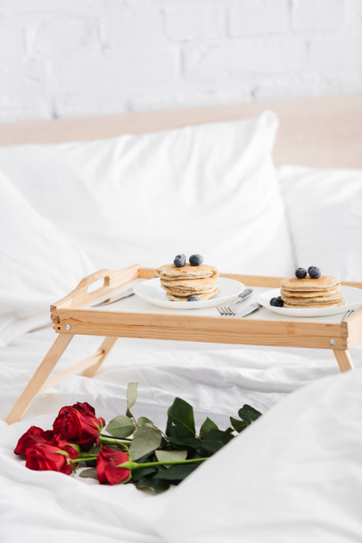 白い寝具に赤いバラの近くにおいしいパンケーキとブルーベリーの木のトレイ、バレンタインデーのコンセプト - 写真・画像