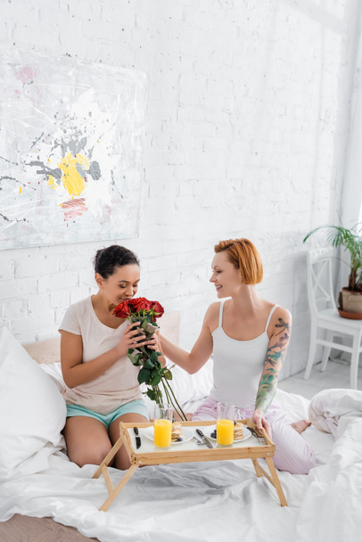 Afro-Amerikan kadın kızıl saçlı lezbiyen kız arkadaşının yanında kırmızı güller kokluyor ve yatakta kahvaltı ediyor. - Fotoğraf, Görsel
