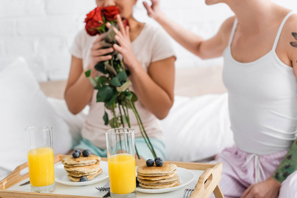 abgeschnittene Ansicht einer lesbischen Frau, die rote Rosen in der Nähe von Freundin und Frühstück im Bett riecht - Foto, Bild