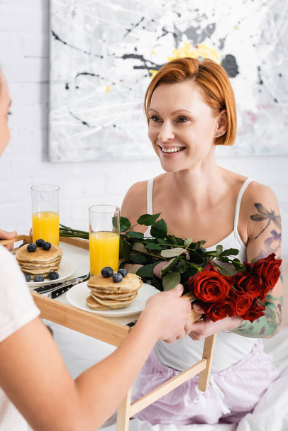 Lesbo nainen tilalla tarjotin aamiaisella ja punaisia ruusuja lähellä onnellinen punapää tyttöystävä - Valokuva, kuva
