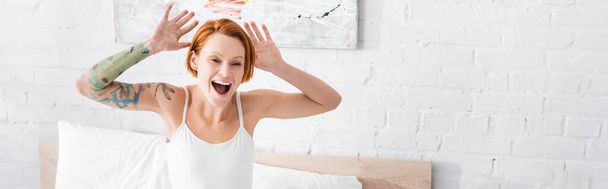excited tattooed woman gesturing with raised hands and shouting in bedroom, banner - Foto, imagen