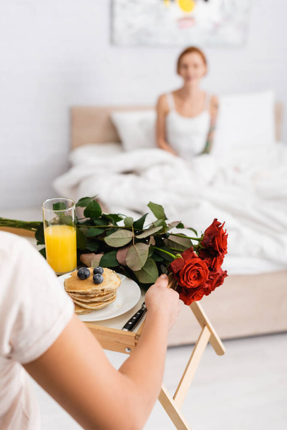 woman holding tray with red roses and breakfast near lesbian girlfriend in bed on blurred background - Photo, Image