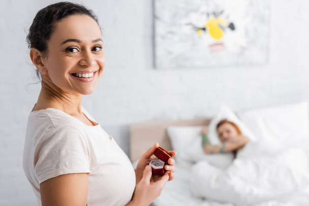 happy african american woman holding jewelry box near lesbian woman lying in bed on blurred background - Фото, изображение