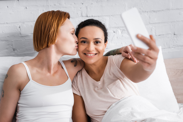 pelirroja lesbiana mujer besos africana americana novia tomando selfie en smartphone en cama - Foto, Imagen