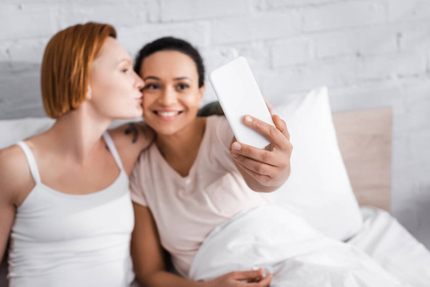 cheerful african american woman taking selfie with redhead lesbian girlfriend kissing her in bed, blurred foreground - Photo, Image