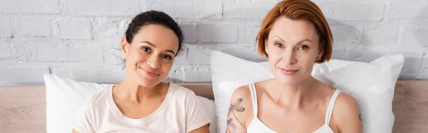 smiling interracial lesbian couple looking at camera in bedroom, banner - Foto, imagen