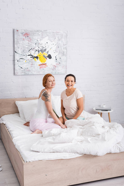 happy interracial lesbian couple smiling at camera in bedroom - Photo, image