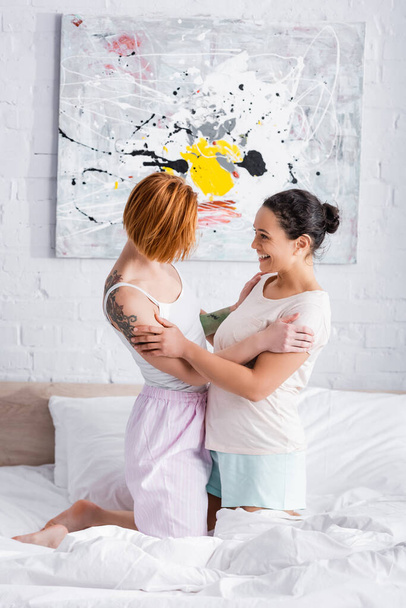side view of redhead lesbian woman and cheerful african american girlfriend hugging and looking at each other in bedroom - Photo, image