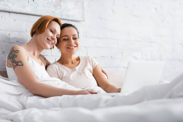 happy interracial lesbian couple watching movie on laptop in bed on blurred foreground - Foto, Imagen