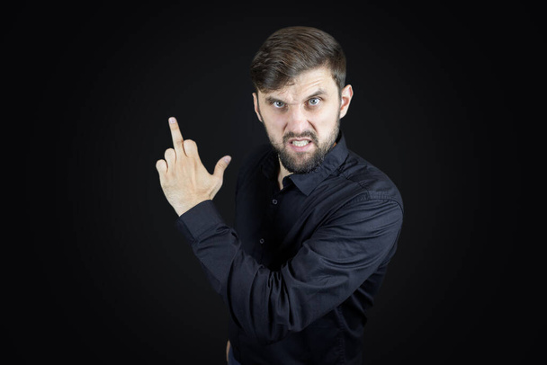 um homem com uma camisa preta e em um fundo preto mostra seus dedos médios faz gestos obscenos - Foto, Imagem