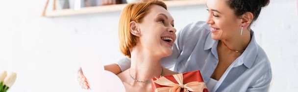 amazed redhead woman with gift box looking at african american lesbian girlfriend, banner - Foto, Bild