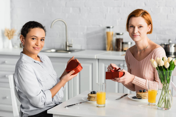 happy interracial lesbian couple looking at camera while holding presents during breakfast on valentines day - Photo, Image