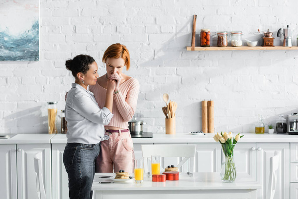lesbian woman kissing hand of african american girlfriend near table with valentines day gifts and breakfast in modern kitchen - Fotoğraf, Görsel
