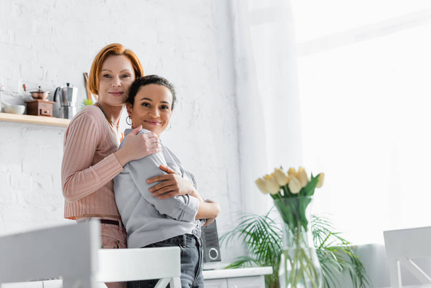 redhead lesbian woman embracing african american girlfriend in kitchen near tulips on blurred foreground - Фото, изображение