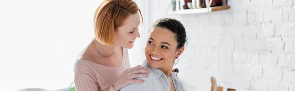 cheerful interracial lesbian couple smiling while looking at each other at home, banner - Photo, image