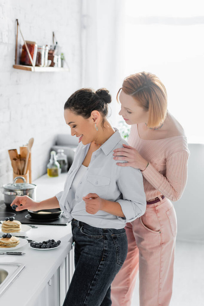 lesbian woman hugging shoulders of happy african american girlfriend serving breakfast in kitchen - Valokuva, kuva