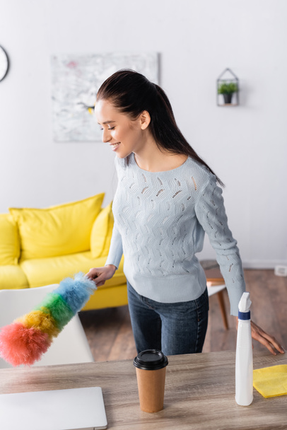 young woman holding dust brush near detergent spray and coffee to go on table - Foto, Imagem