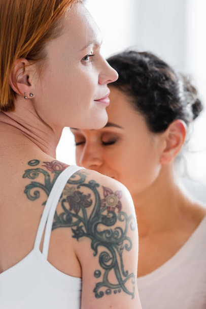 mujer lesbiana tatuada cerca de novia afroamericana con los ojos cerrados sobre fondo borroso - Foto, imagen