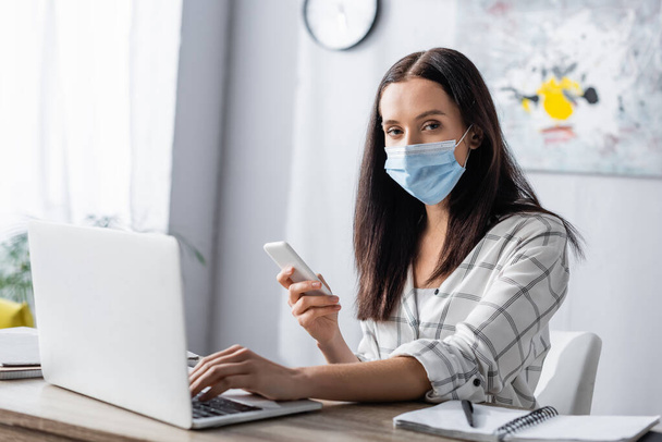 freelancer in medical mask holding smartphone, typing on laptop and looking at camera  - 写真・画像