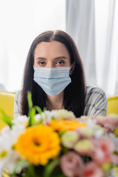 mujer alérgica joven en máscara médica mirando a la cámara cerca de flores en primer plano borrosa - Foto, Imagen