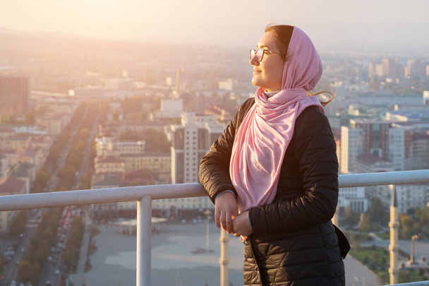 woman with glasses and a pink shawl on the roof of a multi-storey building overlooking the city, sunlight - Фото, изображение