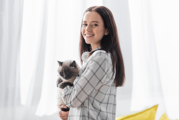 onnellinen nuori nainen katselee kameraa seisoessaan kissan kanssa kotona - Valokuva, kuva
