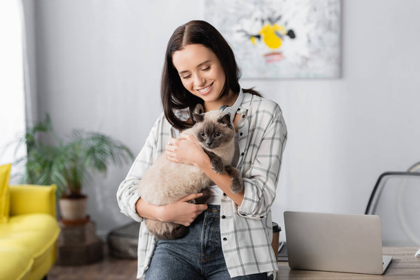 smiling freelancer embracing cat while standing near laptop at home - Foto, imagen