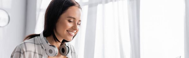 mulher alegre com fones de ouvido sem fio no pescoço sorrindo em casa, banner - Foto, Imagem